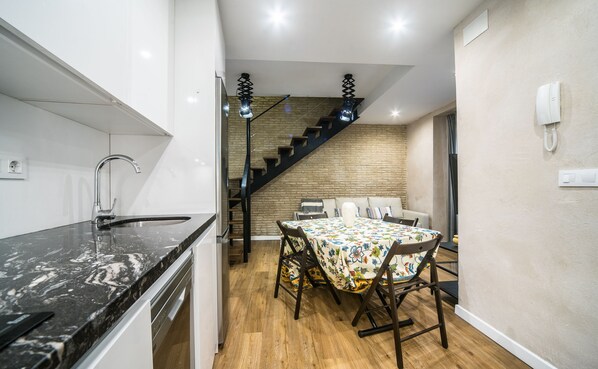 A compact kitchen with a modern sink and dishwasher, a dining area with a floral tablecloth, and a staircase leading to an upper level.
