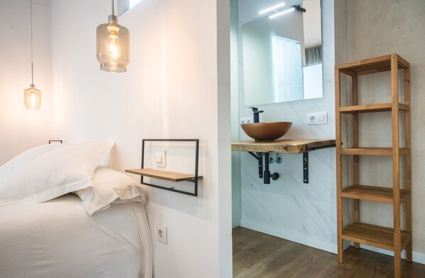 A bedroom with a bed, bedside table, and hanging light fixture. The image also shows a bathroom with a sink, mirror, and wooden shelf.