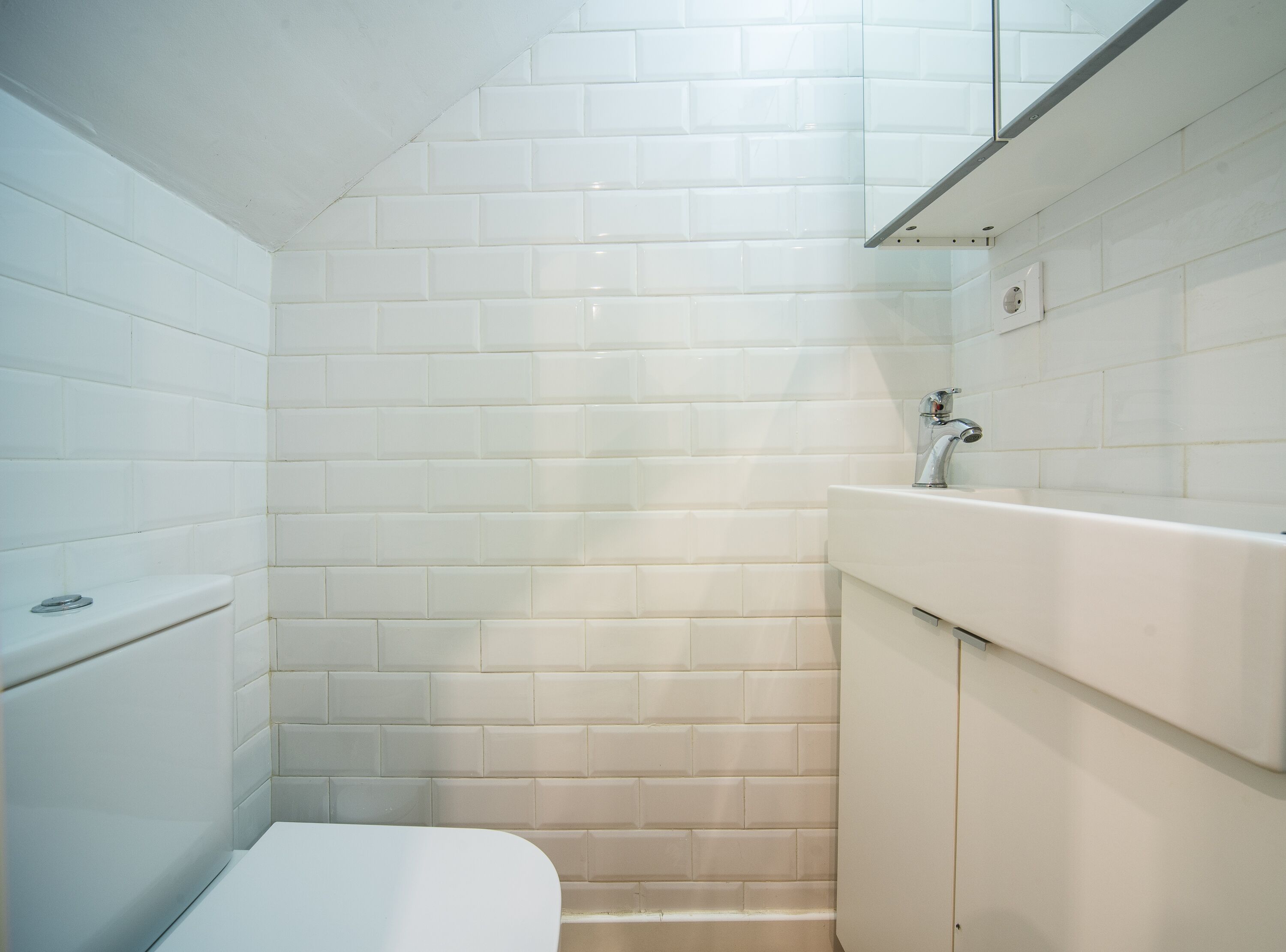 Un baño con paredes de azulejos blancos, un inodoro y un lavamanos con un espejo encima.