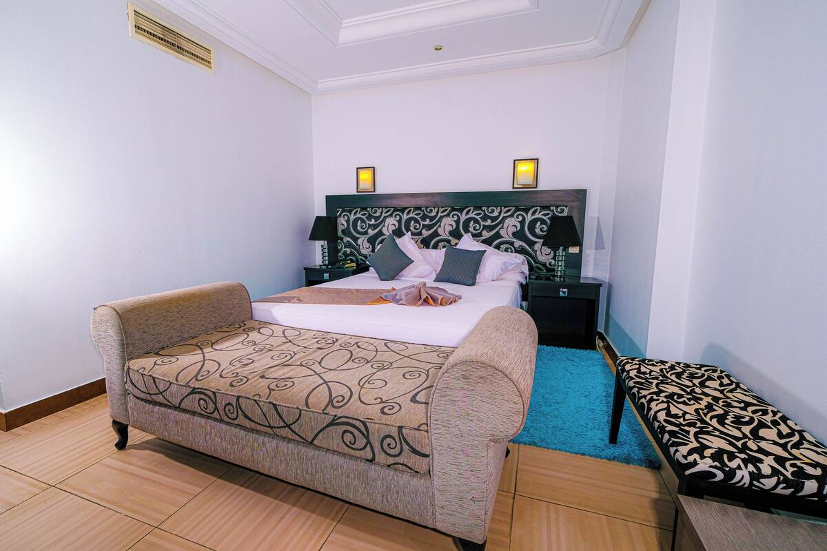Un dormitorio con una cama, un sofá, un banco, una mesita de noche y un calentador en la pared.
