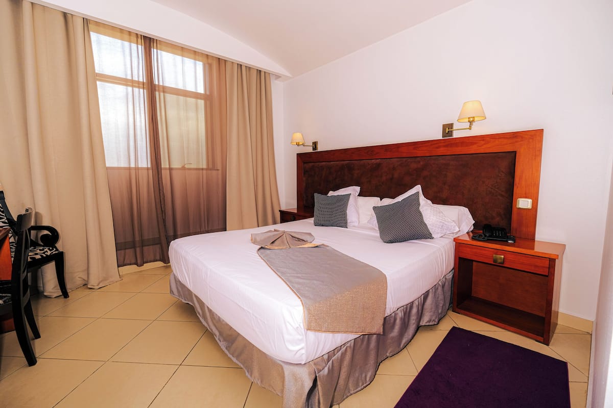 Habitación de hotel con una cama grande, una mesita de noche con lámpara, una silla y una ventana con cortinas.