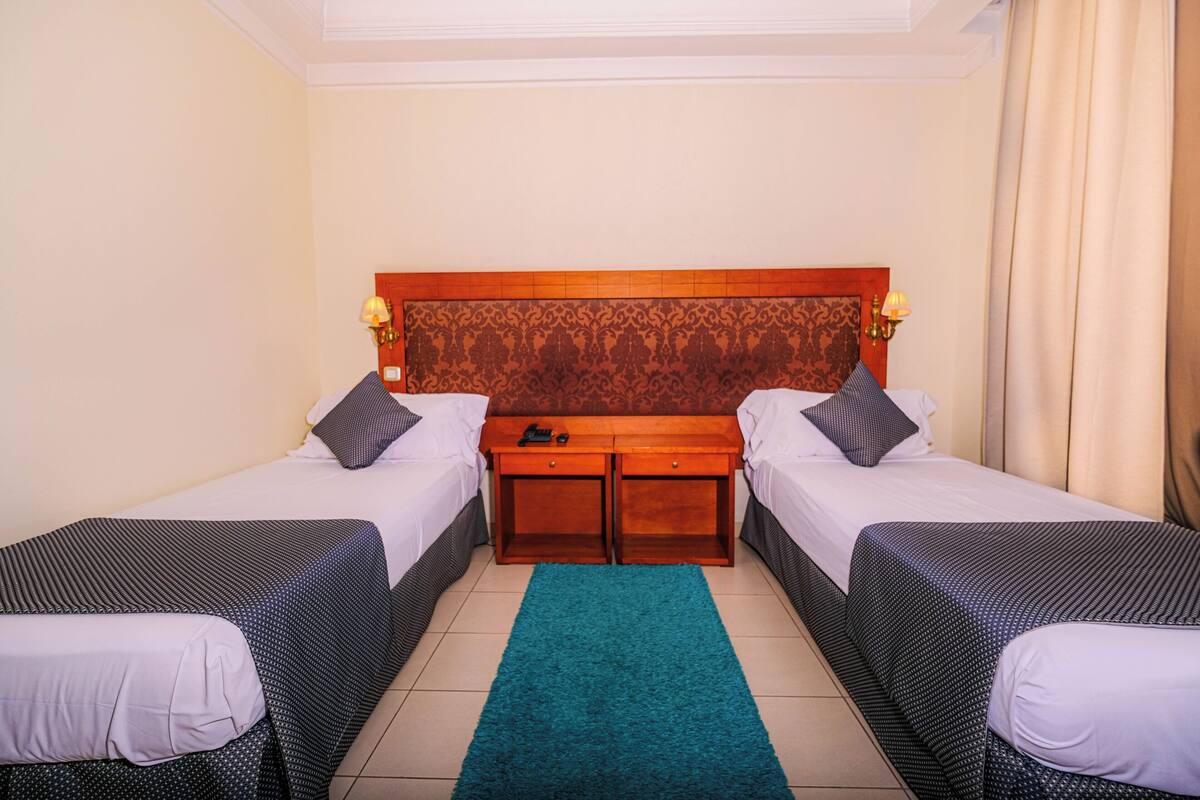 Habitación de hotel con dos camas individuales, un cabecero de madera, una mesita con lámpara y una alfombra color aguamarina.