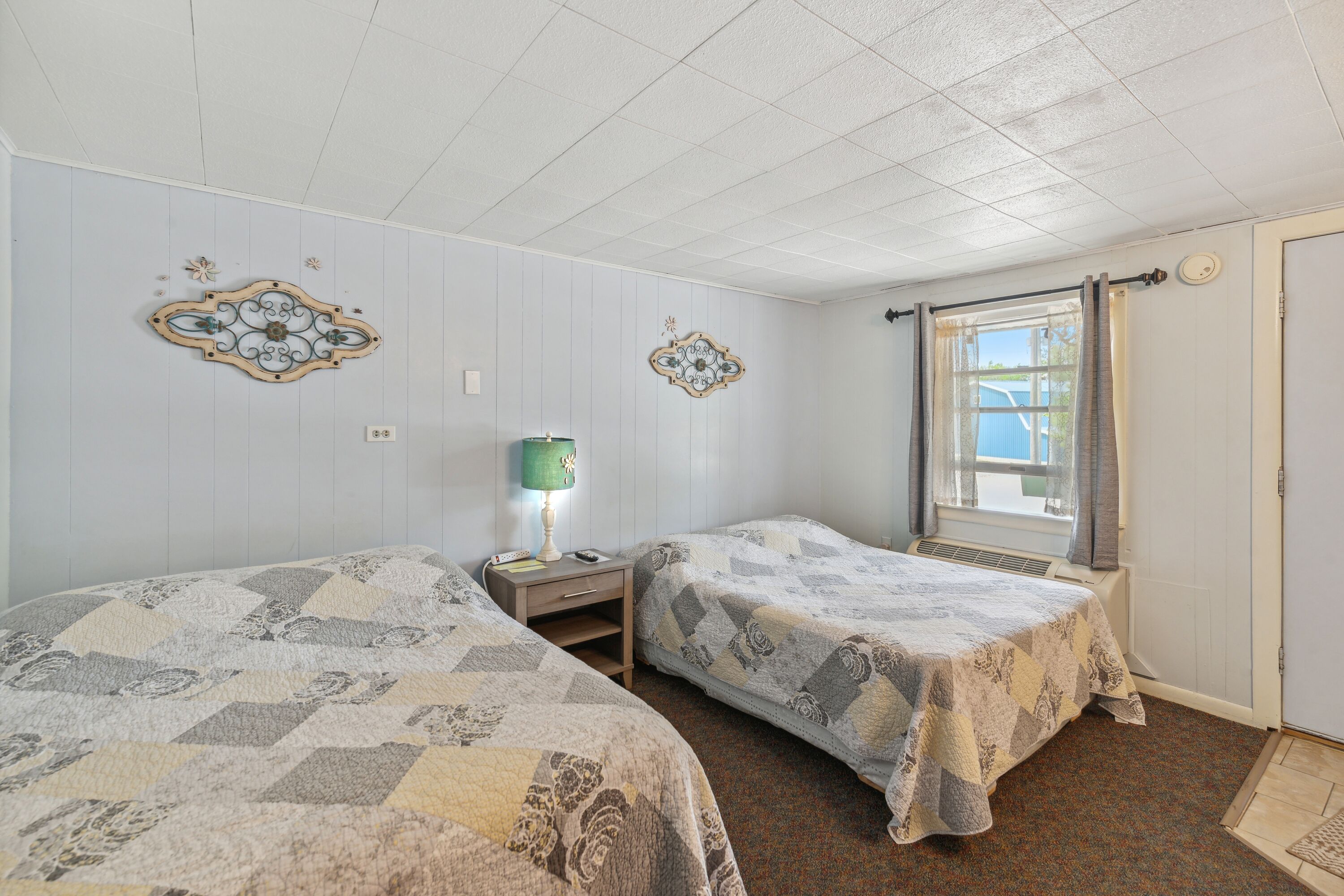 Una habitación con dos camas, una mesita de noche con lámpara, decoración en la pared y una ventana con cortinas.