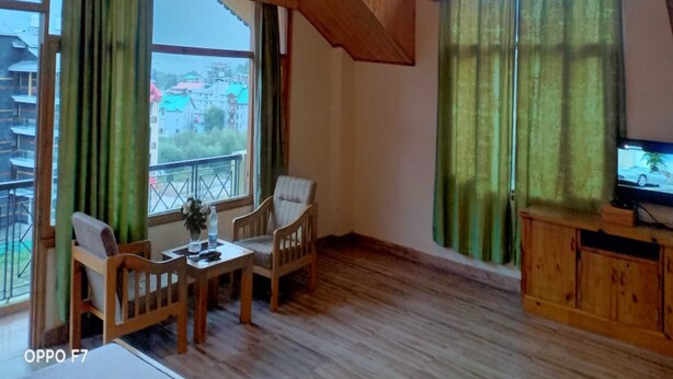 Una habitación acogedora con vigas de madera en el techo, un comedor con sillas y una mesa, un televisor sobre un mueble de madera y ventanas grandes con cortinas verdes.
