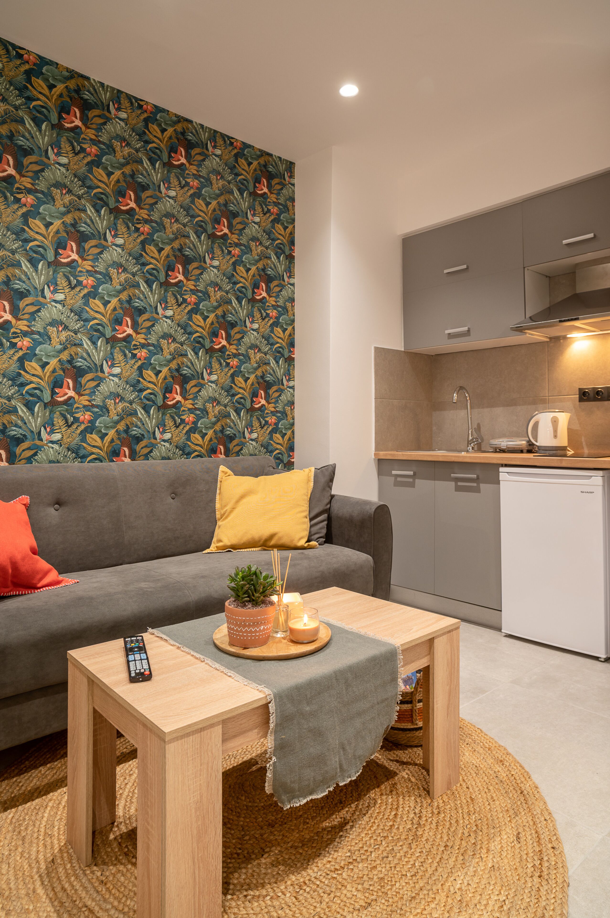 A modern living room with a grey sofa, a small wooden table with a remote control, a potted plant, and a decorative wall.