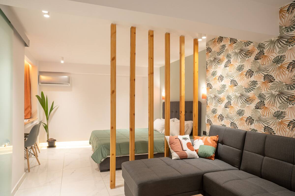 A modern hotel room with a sofa, a bed with green bedding, a desk with a chair, and a potted plant.