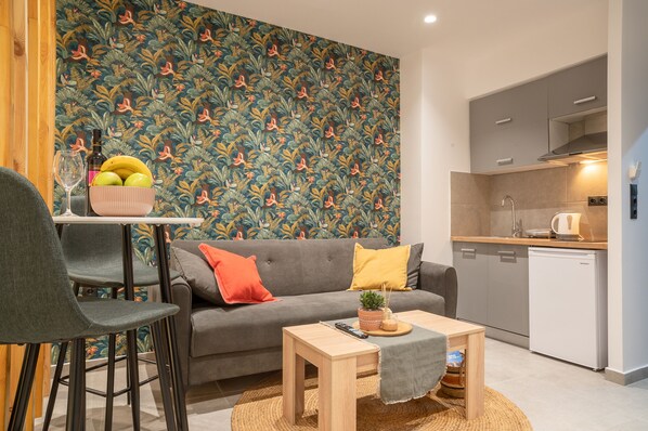 A compact living space with a grey sofa, a small dining table, and a kitchen area with a dishwasher.