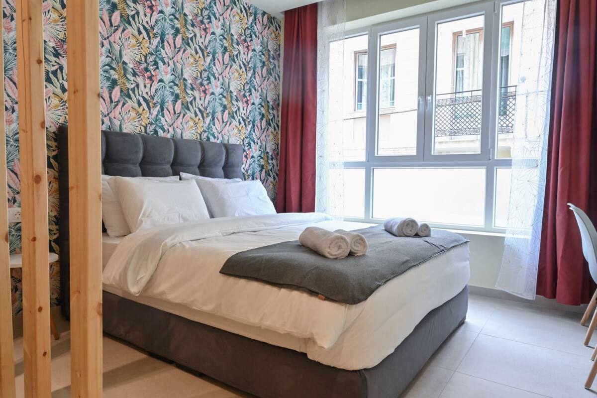 A neatly made bed with white and grey bedding, a floral wallpaper, and a window with red curtains.