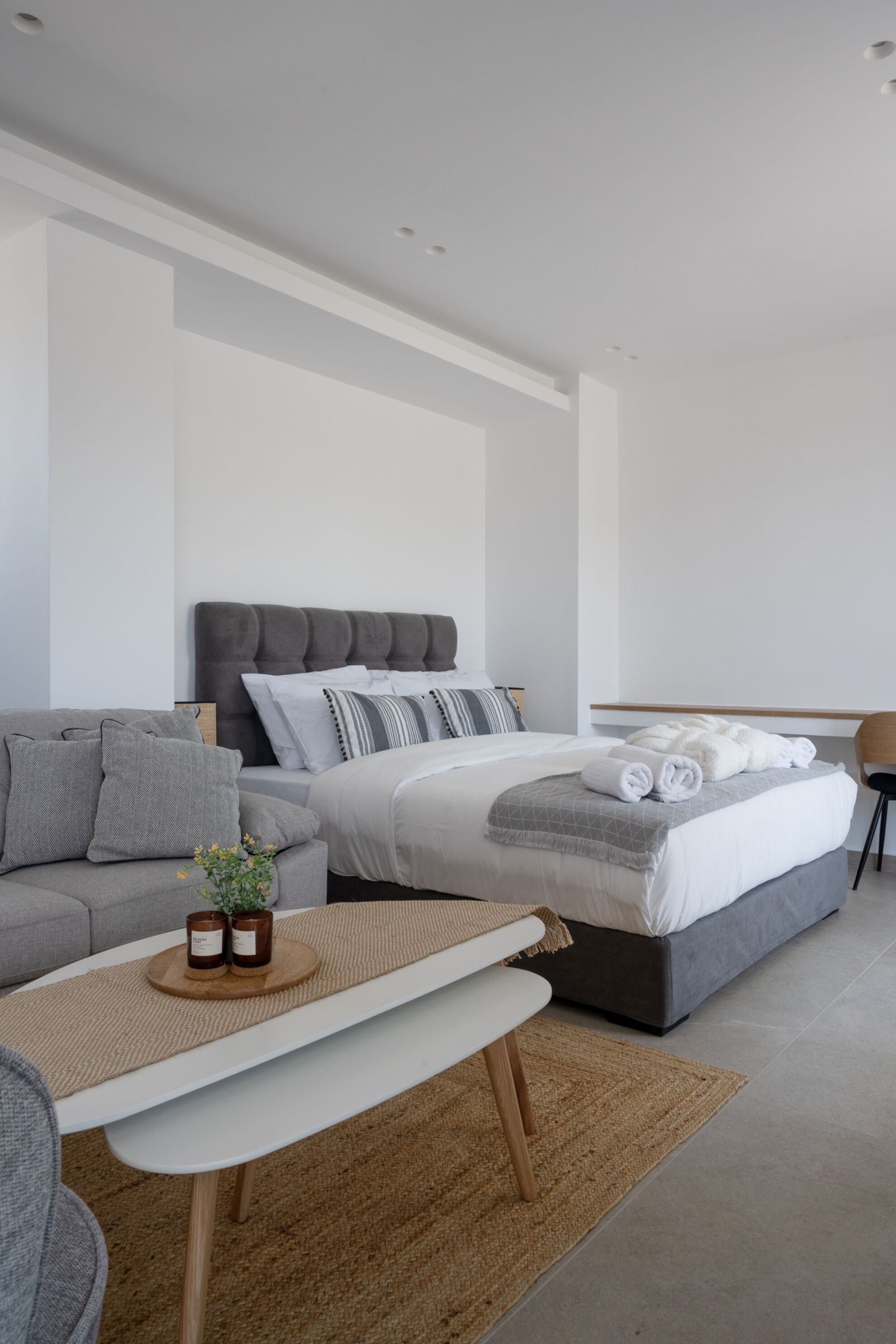 A modern bedroom with a grey sofa, a bed with grey bedding, a small table with candles, and a chair.