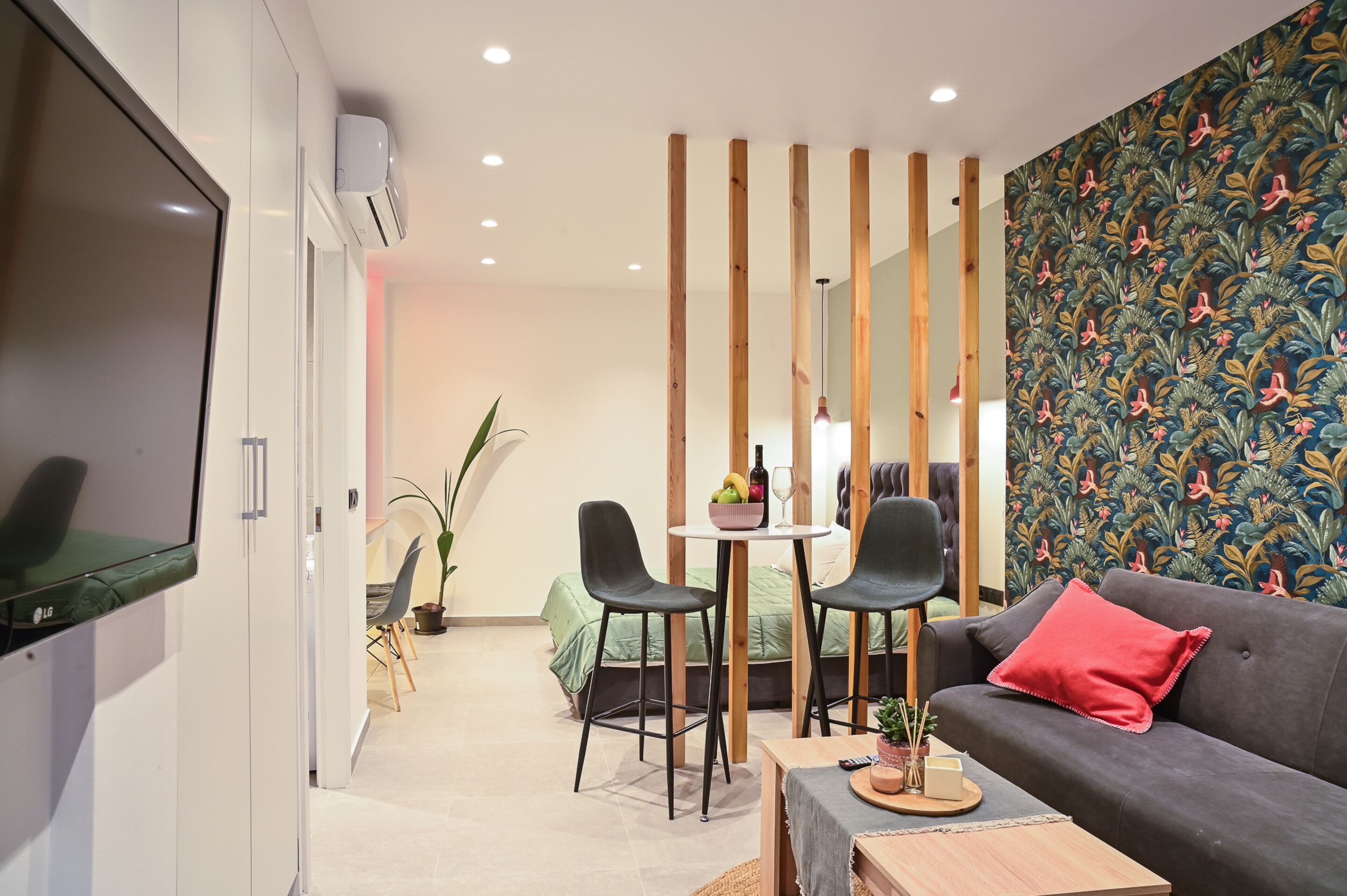 A modern living room with a flat-screen TV, a sofa with a red pillow, a dining area with chairs and a table, and a patterned wallpaper.
