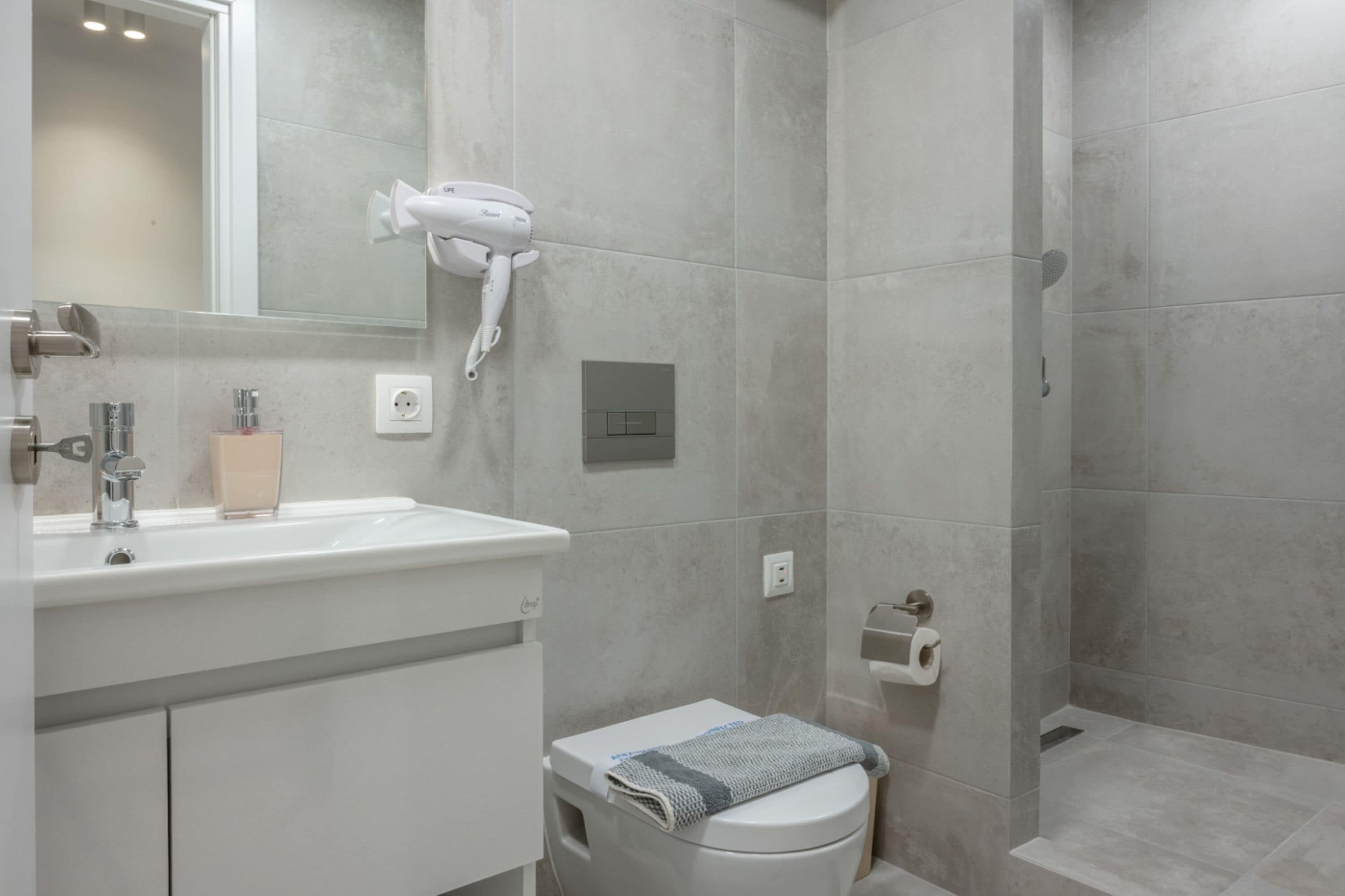 A modern bathroom with a white sink, mirror, and toilet. There is a hairdryer mounted on the wall and a towel on the toilet.