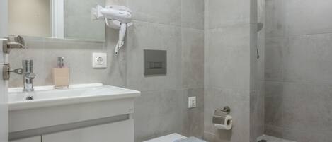 A modern bathroom with a white sink, mirror, and toilet. There is a hairdryer mounted on the wall and a towel on the toilet.