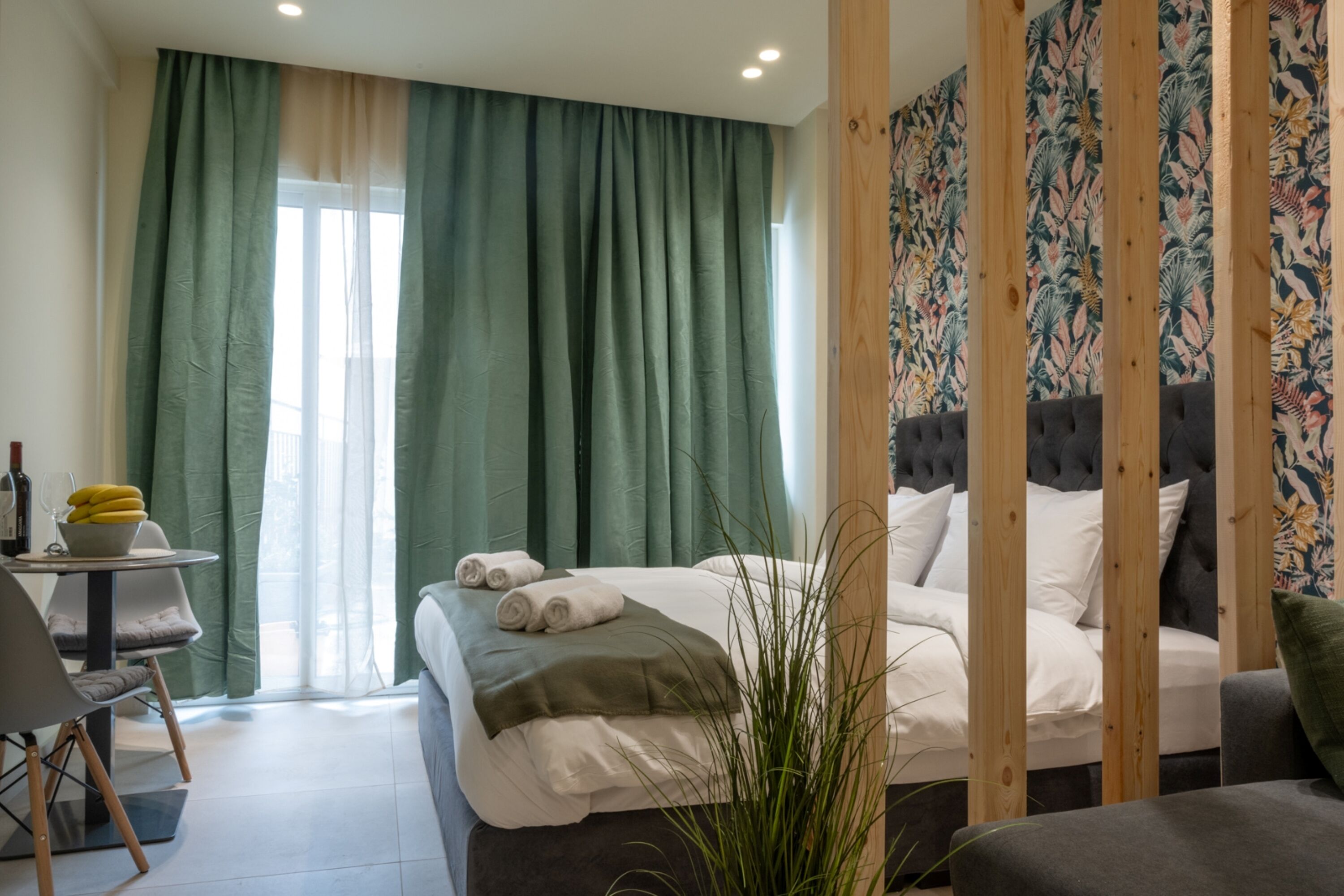 A hotel room with a bed, a small table with a bowl of fruit and wine, a chair, and a potted plant.