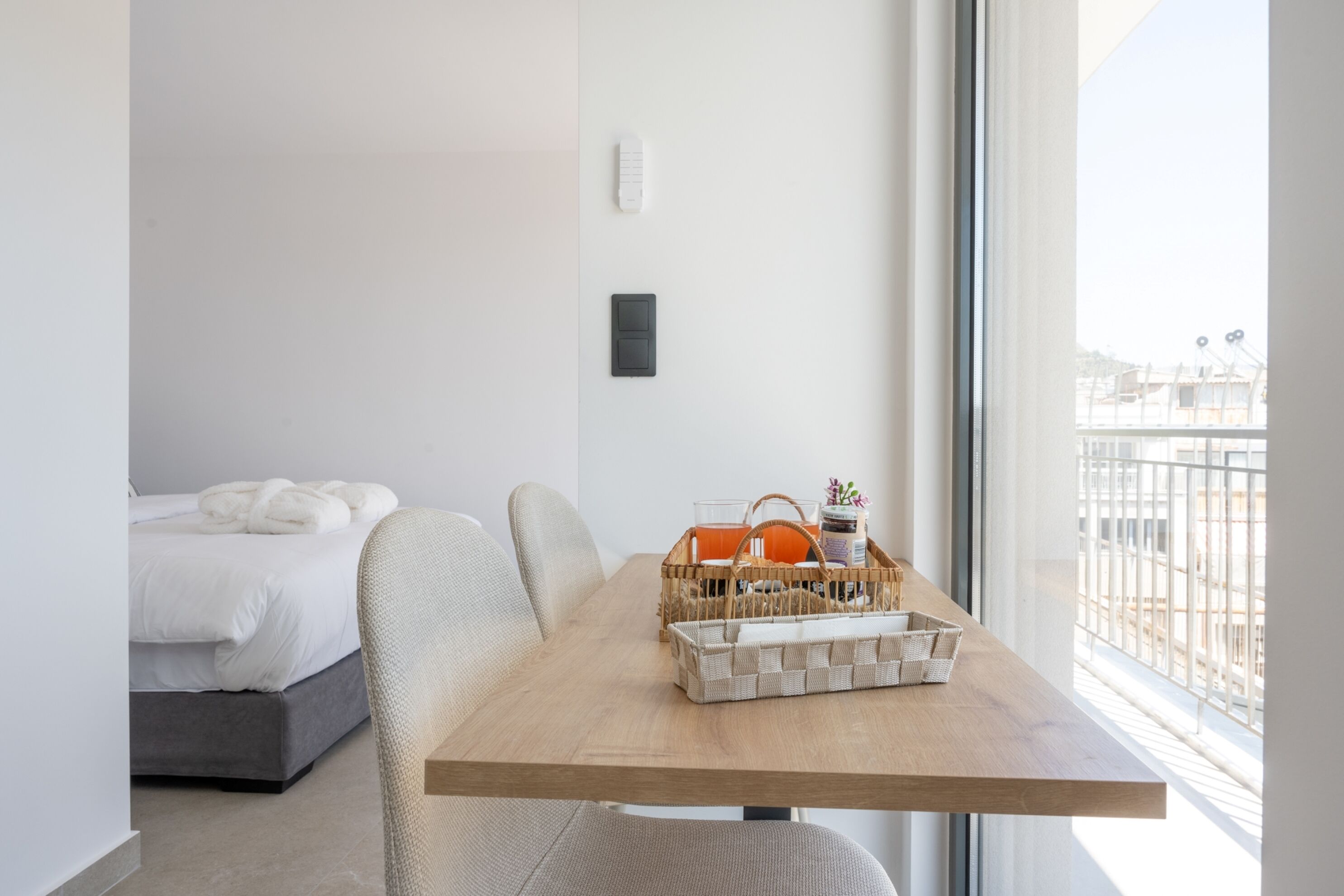 A modern hotel room with a bed, a dining table with a basket of items, and a view of the city through the window.