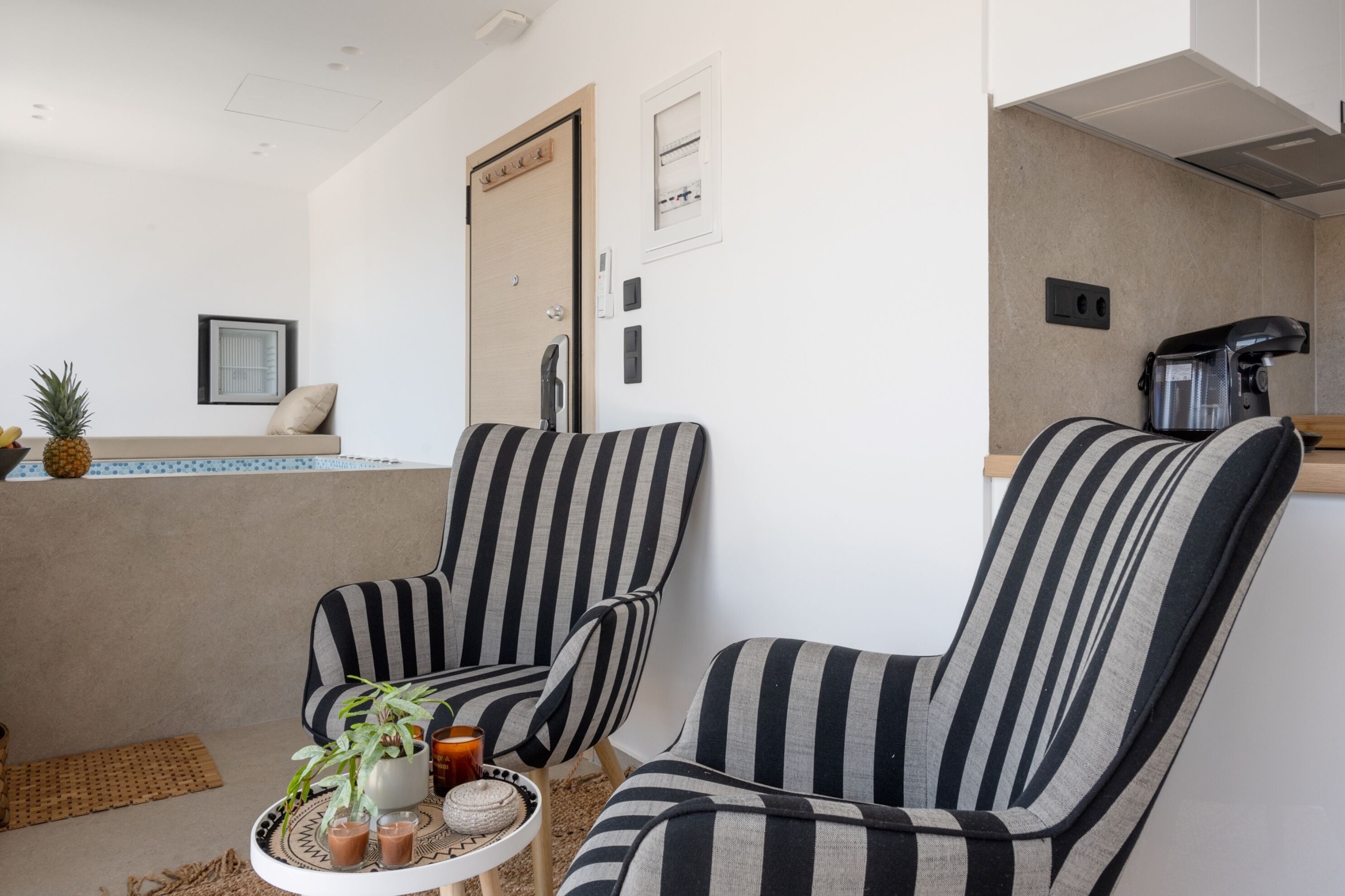 A room with two striped armchairs, a small table with drinks and a plant, a door, and a kitchenette area.