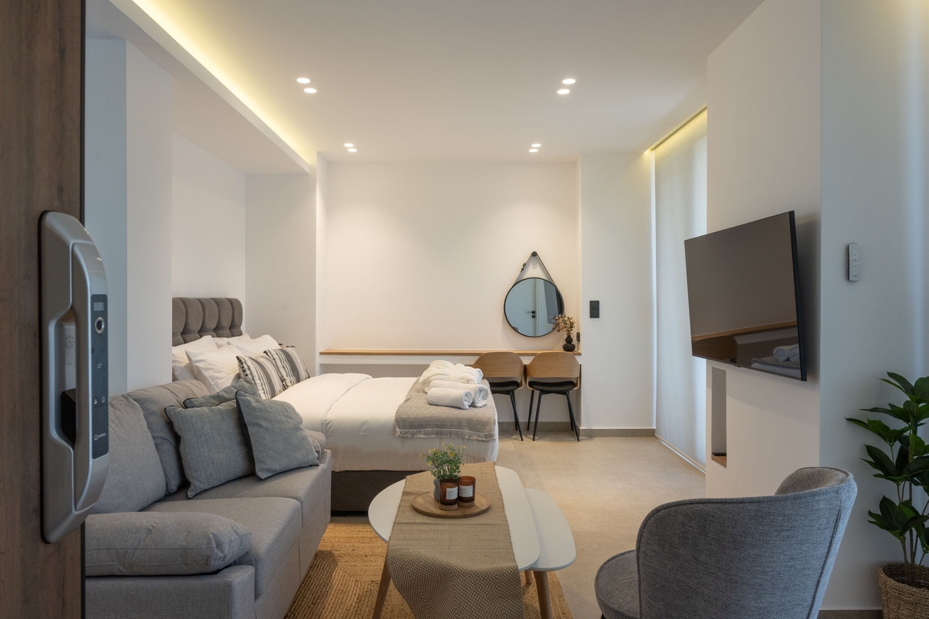 A modern hotel room with a bed, a sofa, a desk, a mirror, and a TV.