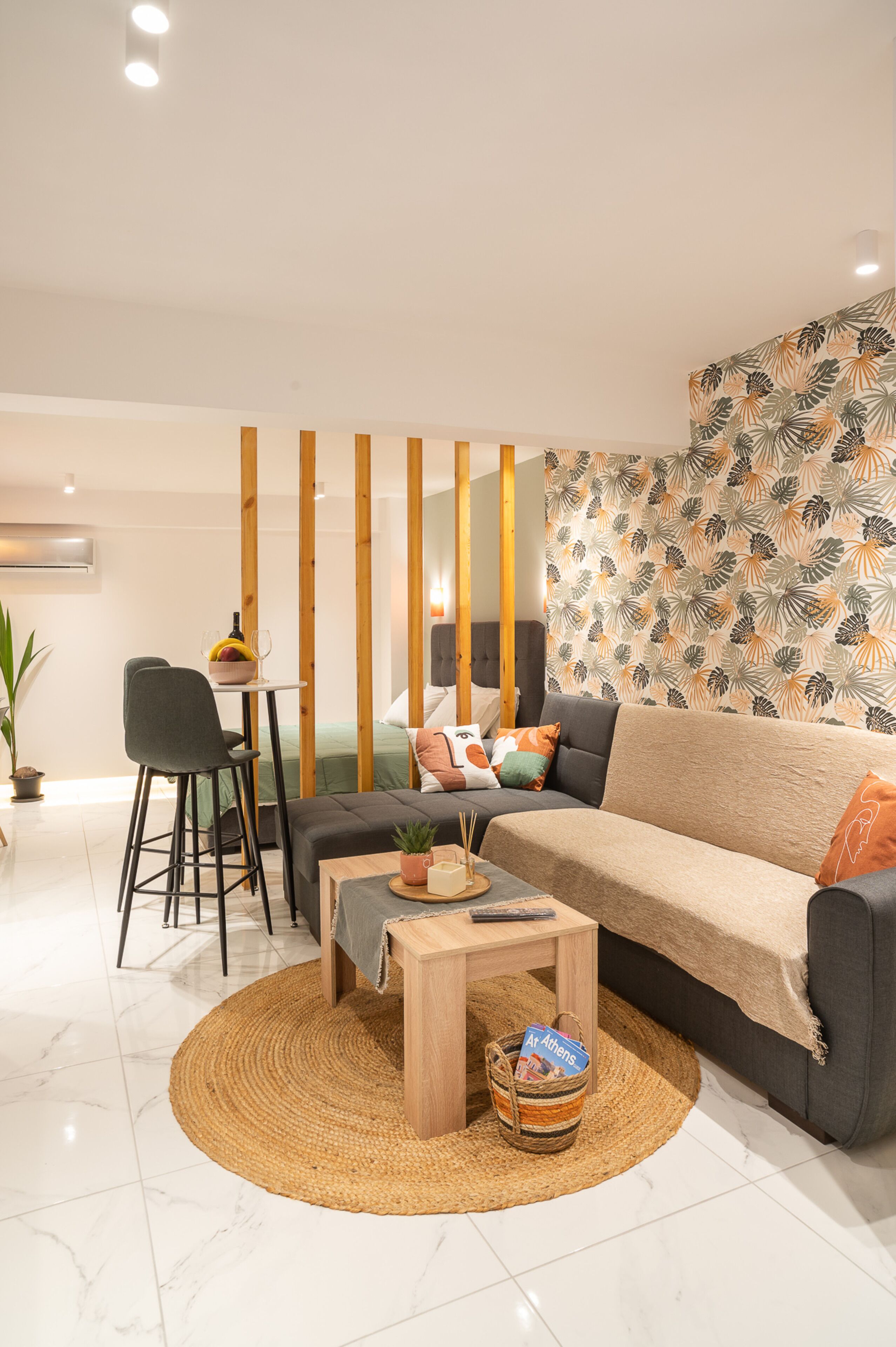 Una sala moderna con sofá, mesa de centro, comedor con una silla y una mesita, y una pared decorativa con motivo de hojas.