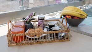 A breakfast setup with a wicker basket containing a drink, pastries, and a bowl of fruit on a balcony with a pool view. - Heart of Athens Apartments (Athens)