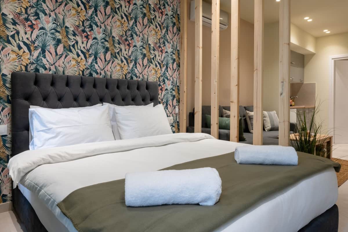 A neatly made bed with white linens and a green throw, a tufted headboard, and a tropical leaf-patterned wall.