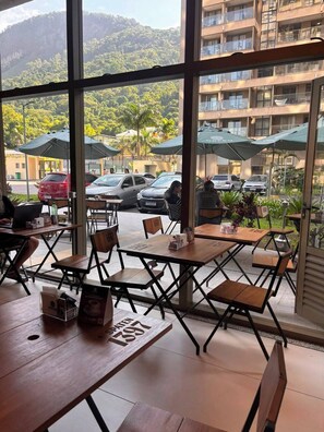 An indoor seating area with tables and chairs, large windows offering a view of parked cars and a building with balconies.