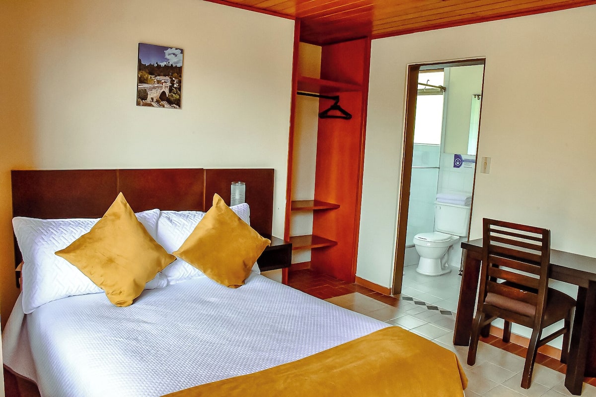Quarto de hotel com uma cama, cabeceira de madeira, uma mesa pequena com uma cadeira, banheiro visível por uma porta aberta e um porta-roupas fixado na parede.