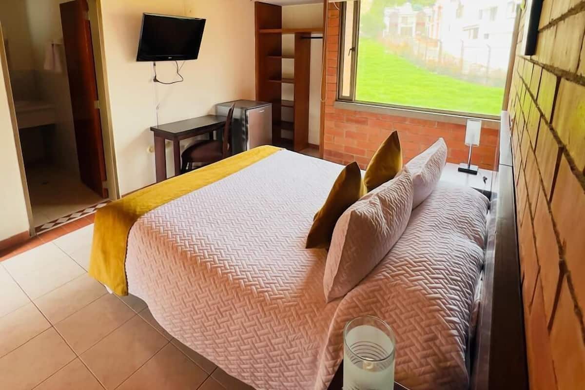Quarto com teto de madeira, cama com colcha estampada, mesa de cabeceira com abajur e TV na parede.