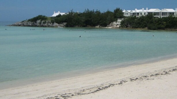 Uma praia com um edifício branco numa ilha pequena, água turquesa cristalina e uma costa arenosa.