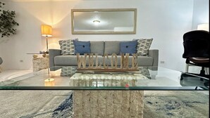 Uma sala de estar moderna com uma mesa de centro de vidro, um sofá cinza com almofadas estampadas e um letreiro com a palavra 'WELCOME'.