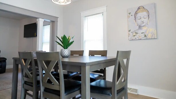 Een eethoek met een tafel, stoelen, een potplant, een Boeddhabeeld en een televisie op de achtergrond.