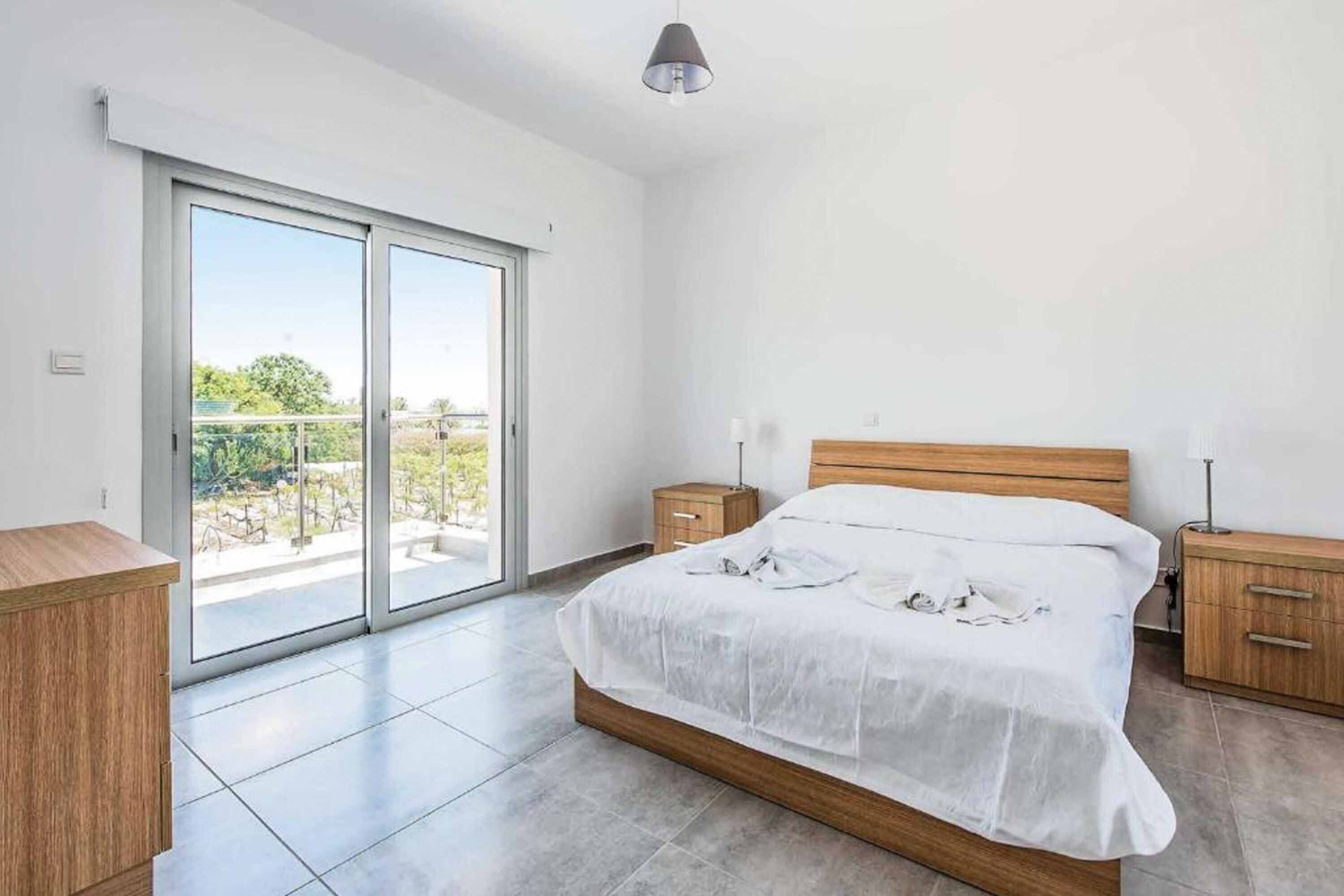 A bedroom with a large bed, a wooden headboard, a nightstand, a bedside table, and a sliding glass door leading to a balcony.