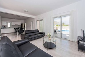 A modern living area with a black sofa, a glass coffee table, and a TV stand. A kitchen is visible in the background, and there is a sliding glass door leading to an outdoor area. - Demitrios Vine - Three Bedroom Villa, Sleeps 6 (Peyia)