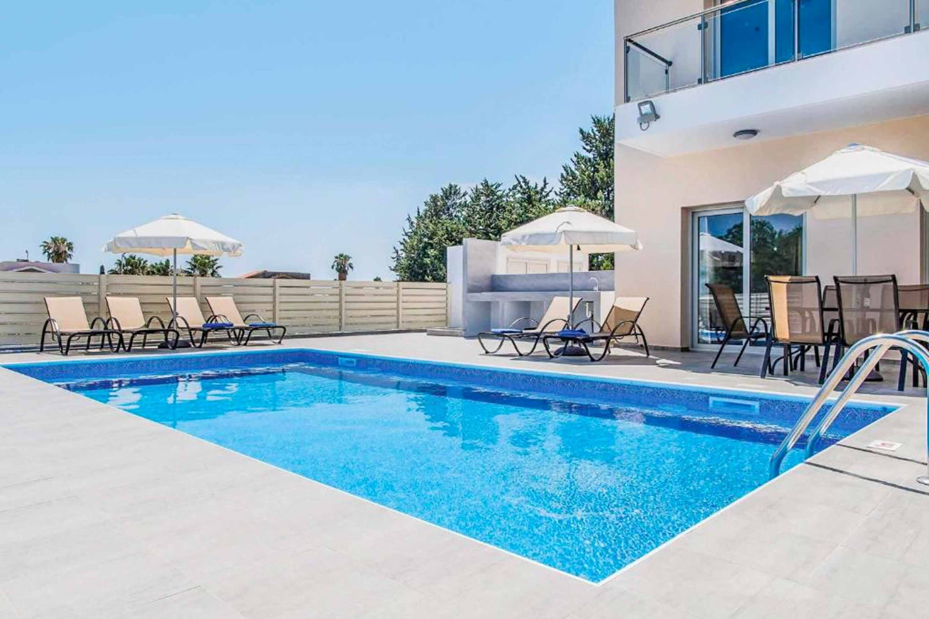 Una piscina con sillas de descanso y sombrillas, una zona de estar techada y un edificio con ventanas amplias.