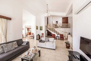 A spacious living area with a sofa, coffee table, TV, and a staircase leading to an upper level. - Villa Theo - Four Bedroom Villa, Sleeps 8 (Peyia)