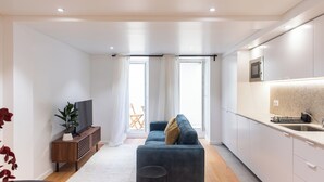 A modern apartment with a living area featuring a sofa, a TV, and a small side table. There is a kitchen with white cabinets and a microwave. - The Haus - Alegria (Lisbon)