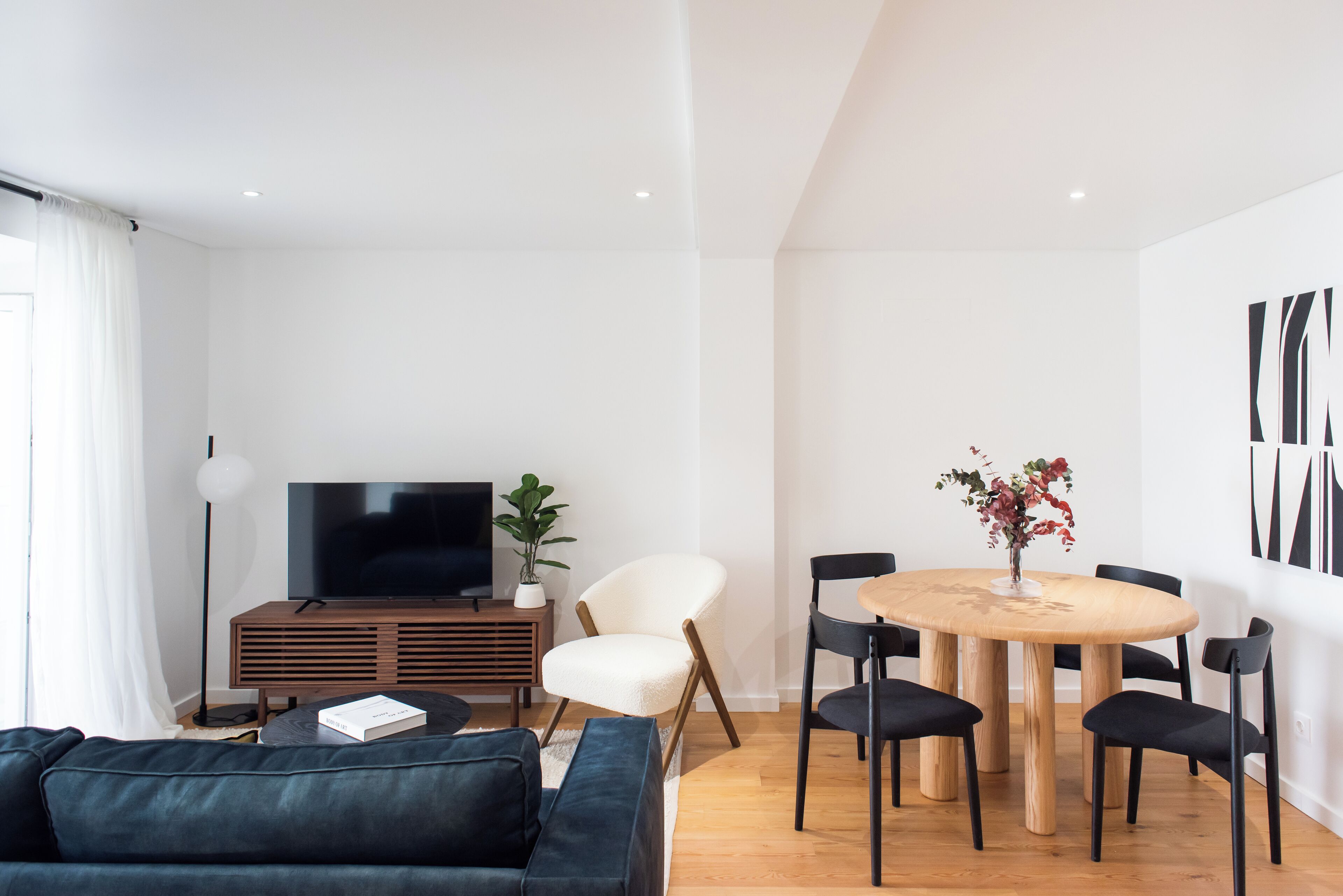 A modern living room with a dark-colored sofa, a wooden TV stand, a white armchair, a round wooden dining table with black chairs, and a potted plant.