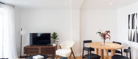 A modern living room with a dark-colored sofa, a wooden TV stand, a white armchair, a round wooden dining table with black chairs, and a potted plant.