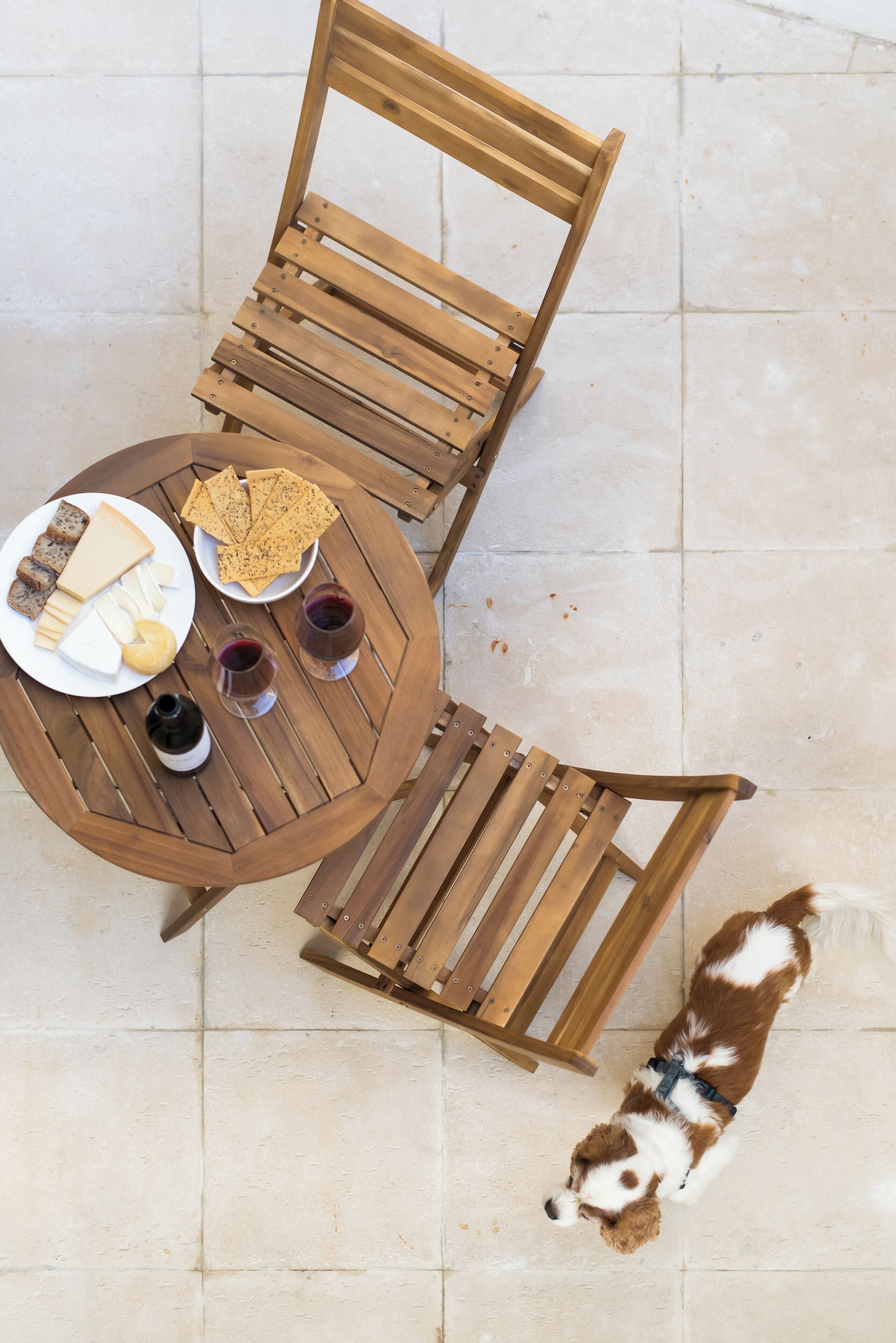 A round wooden table with a plate of cheese, crackers, and wine glasses, surrounded by wooden chairs.