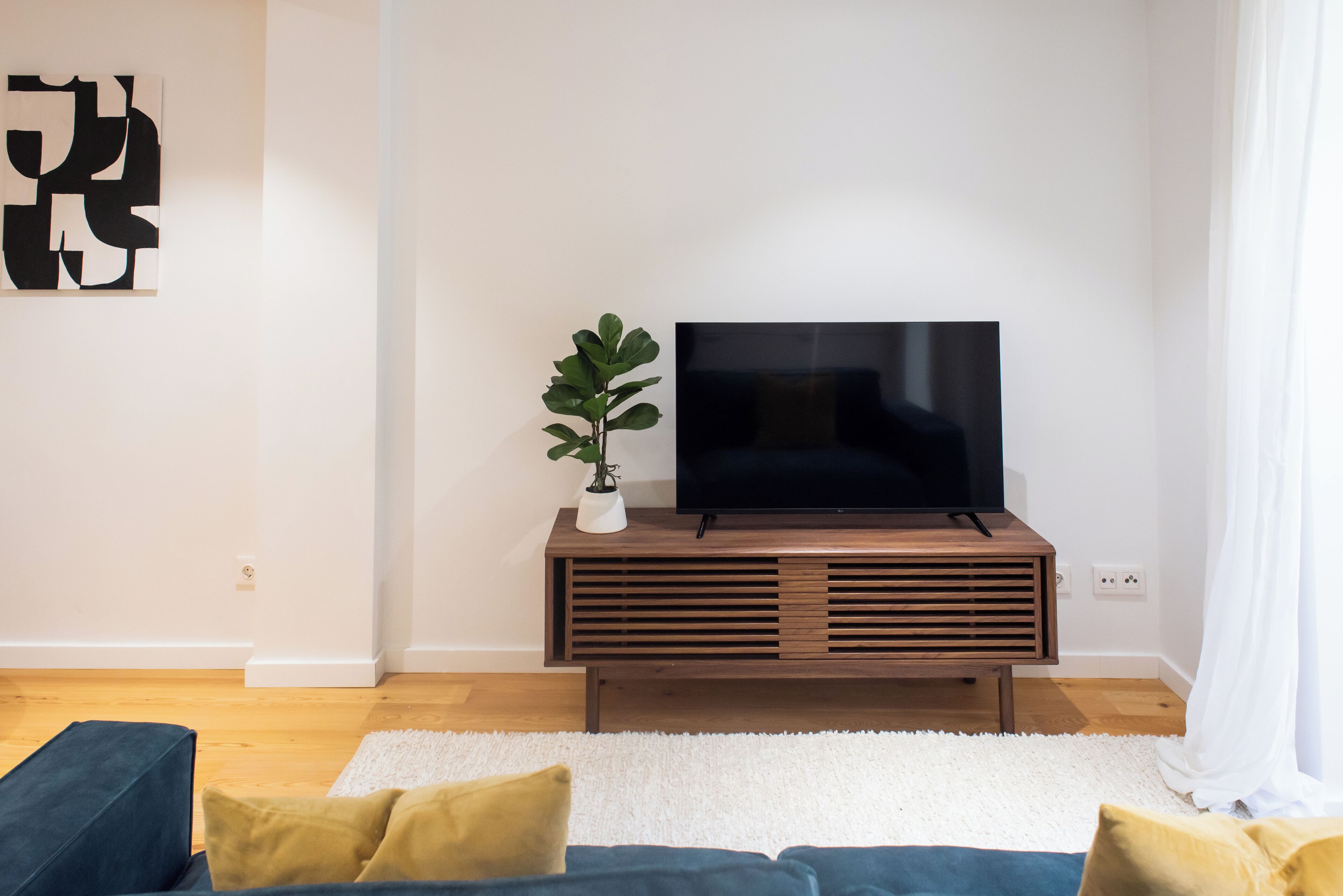 A modern living room with a flat-screen TV on a wooden stand, a potted plant, and a blue sofa with yellow cushions.