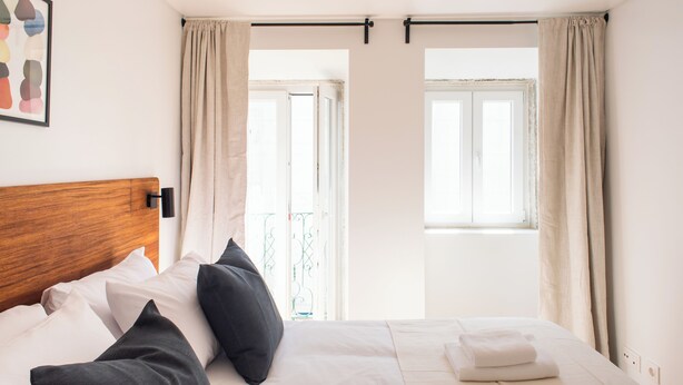 A neatly made bed with white and dark pillows, a wooden headboard, and a window with curtains. - The Haus - Alegria (Lisbon)