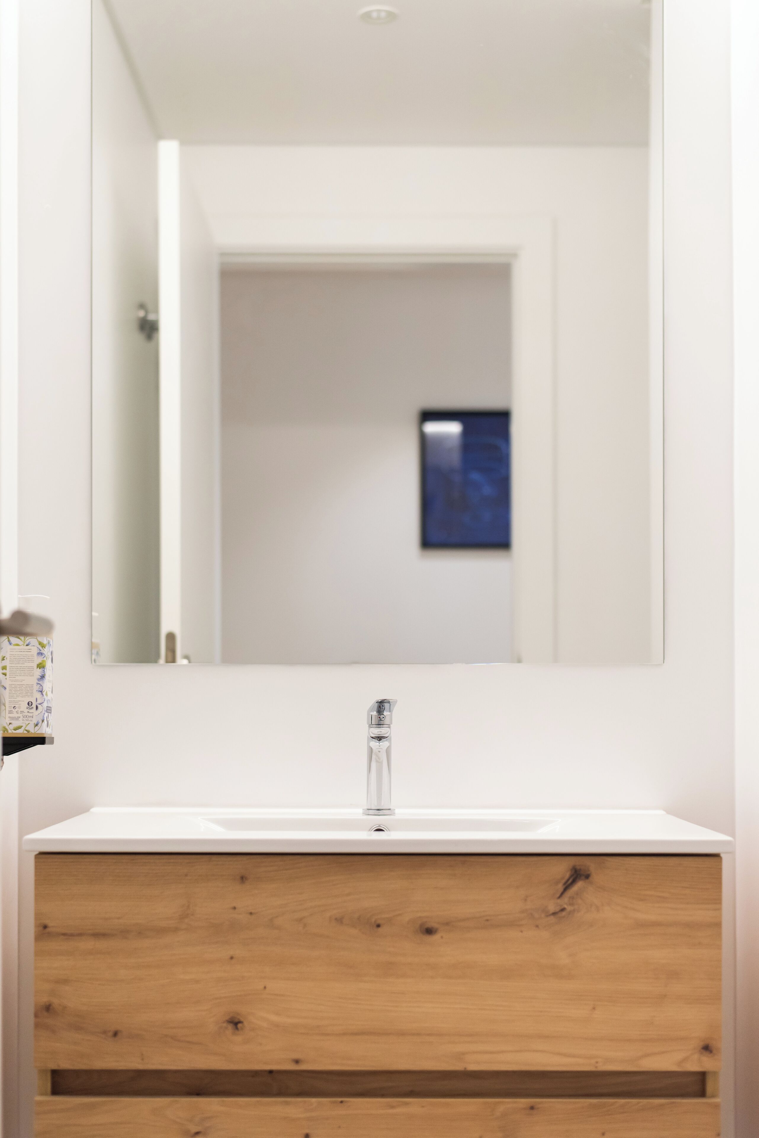 Une salle de bain avec un lavabo blanc, un meuble en bois et un miroir qui reflète une porte et un tableau encadré accroché au mur.