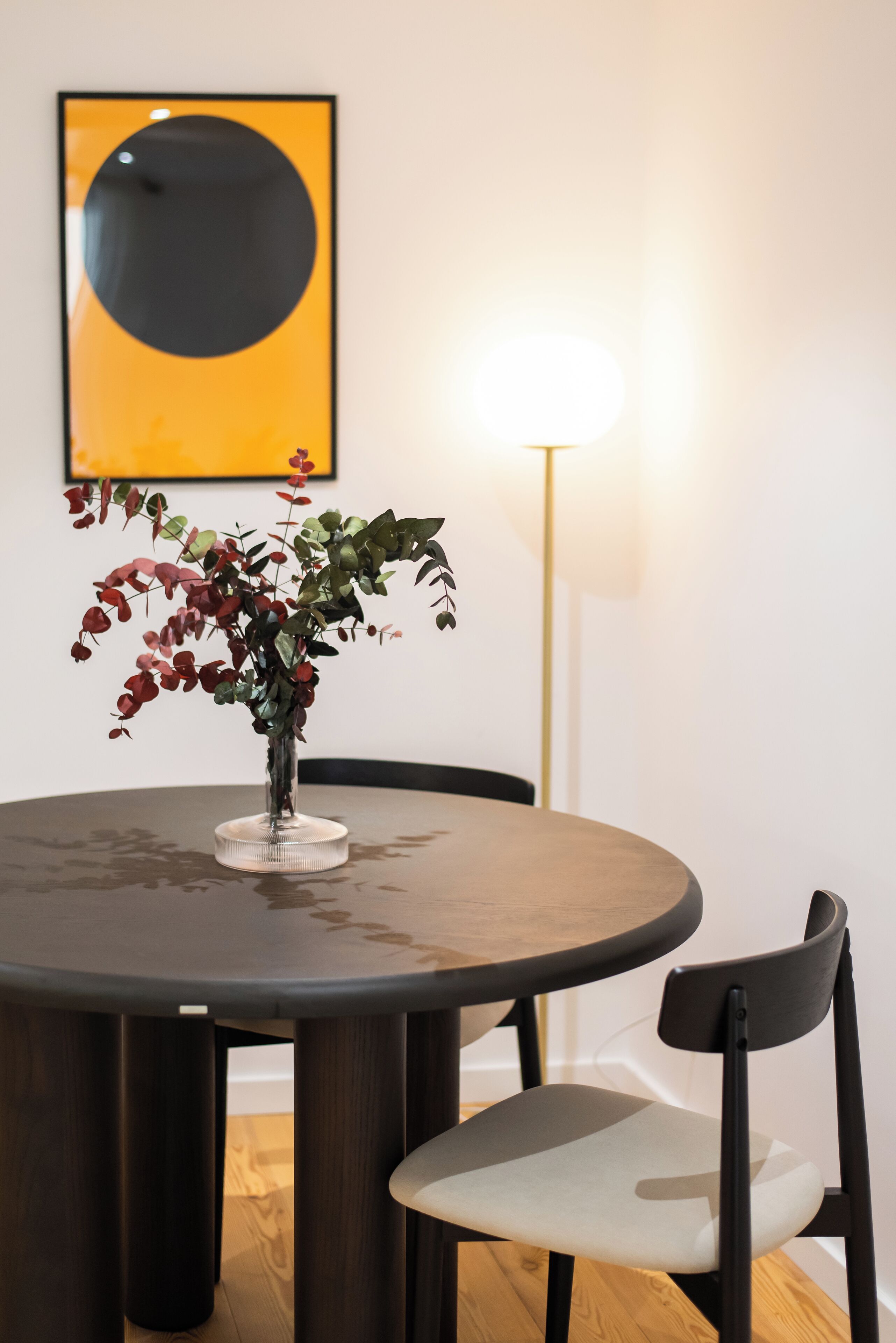 A round dining table with a vase of red-leafed plants, a black chair, and a floor lamp.