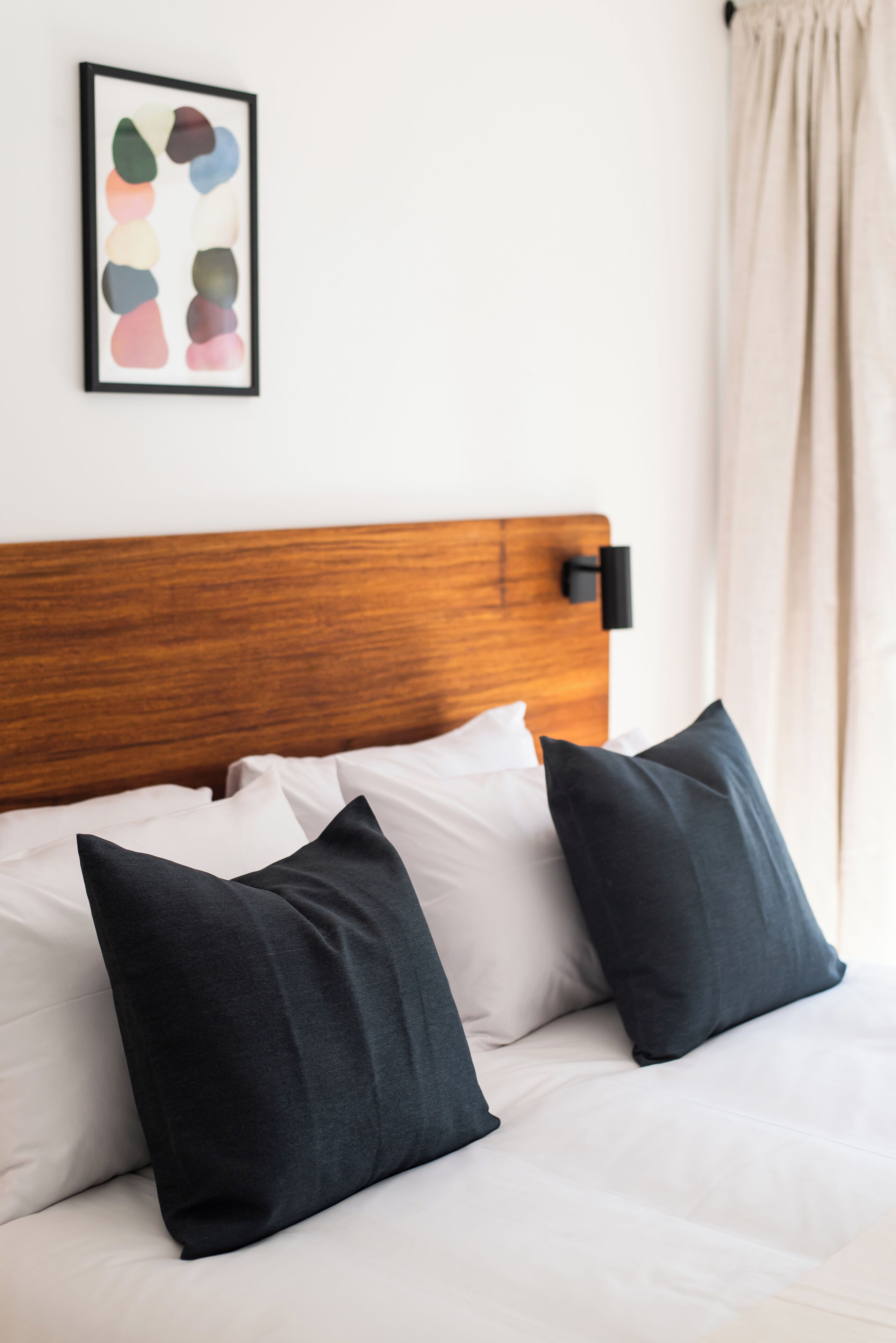 Un lit bien fait, avec des oreillers blancs et foncés, une tête de lit en bois et une œuvre d’art abstraite encadrée accrochée au mur.