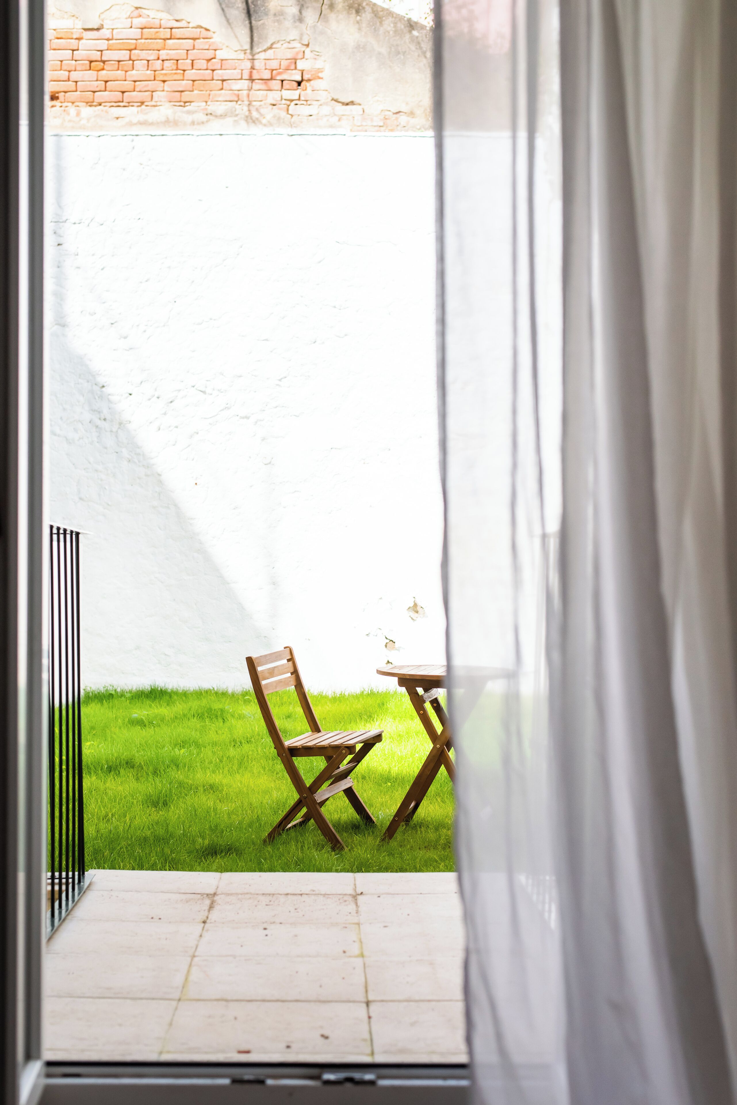 Blick durch ein Fenster mit weißem Vorhang auf einen Innenhof mit Rasenfläche, einem Holzstuhl und einem kleinen Tisch.