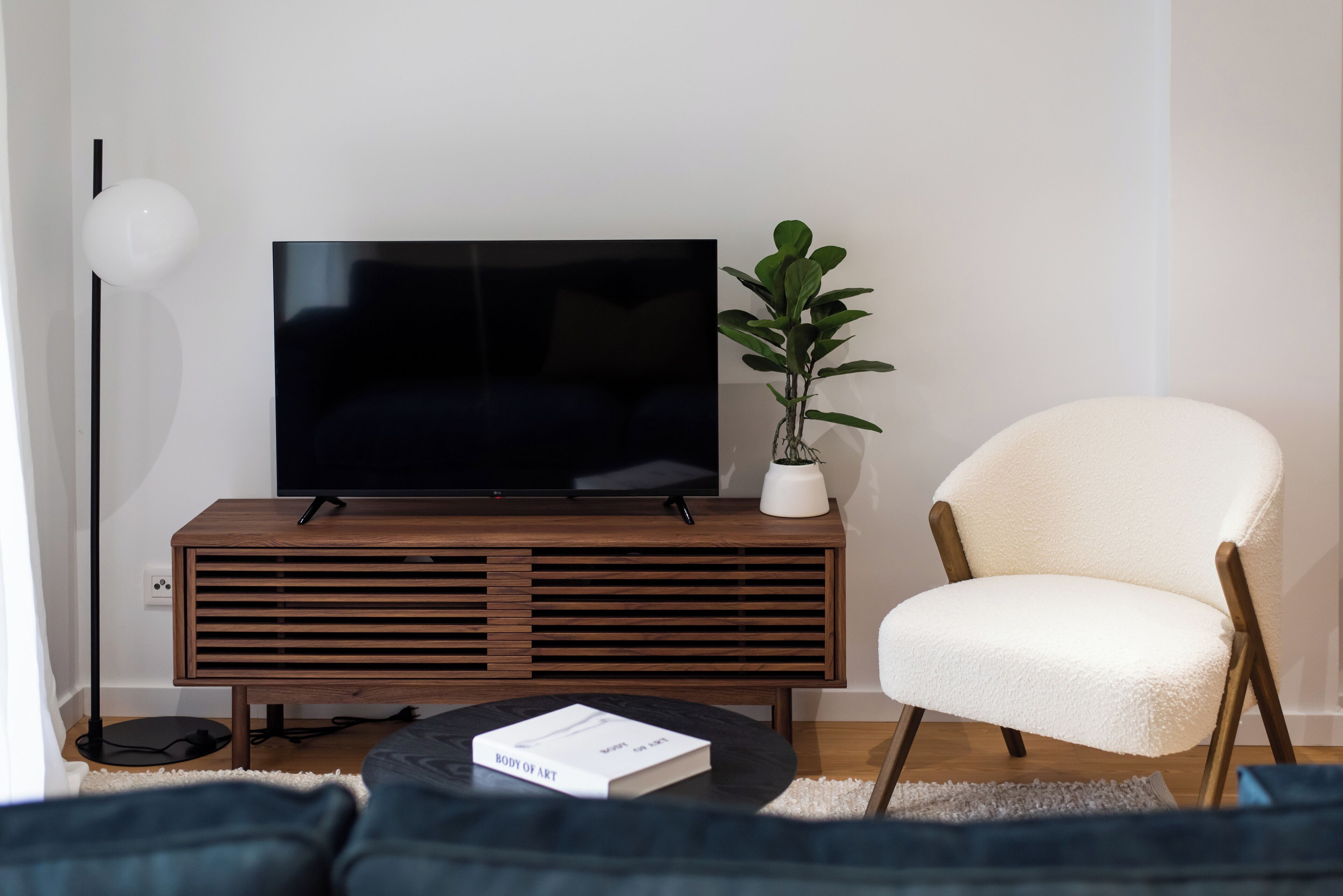 Ein modernes Wohnzimmer mit einem Flachbildfernseher auf einem Holzboden, einem weißen Sessel, einer kleinen Topfpflanze und einem Buch auf einem runden Couchtisch.