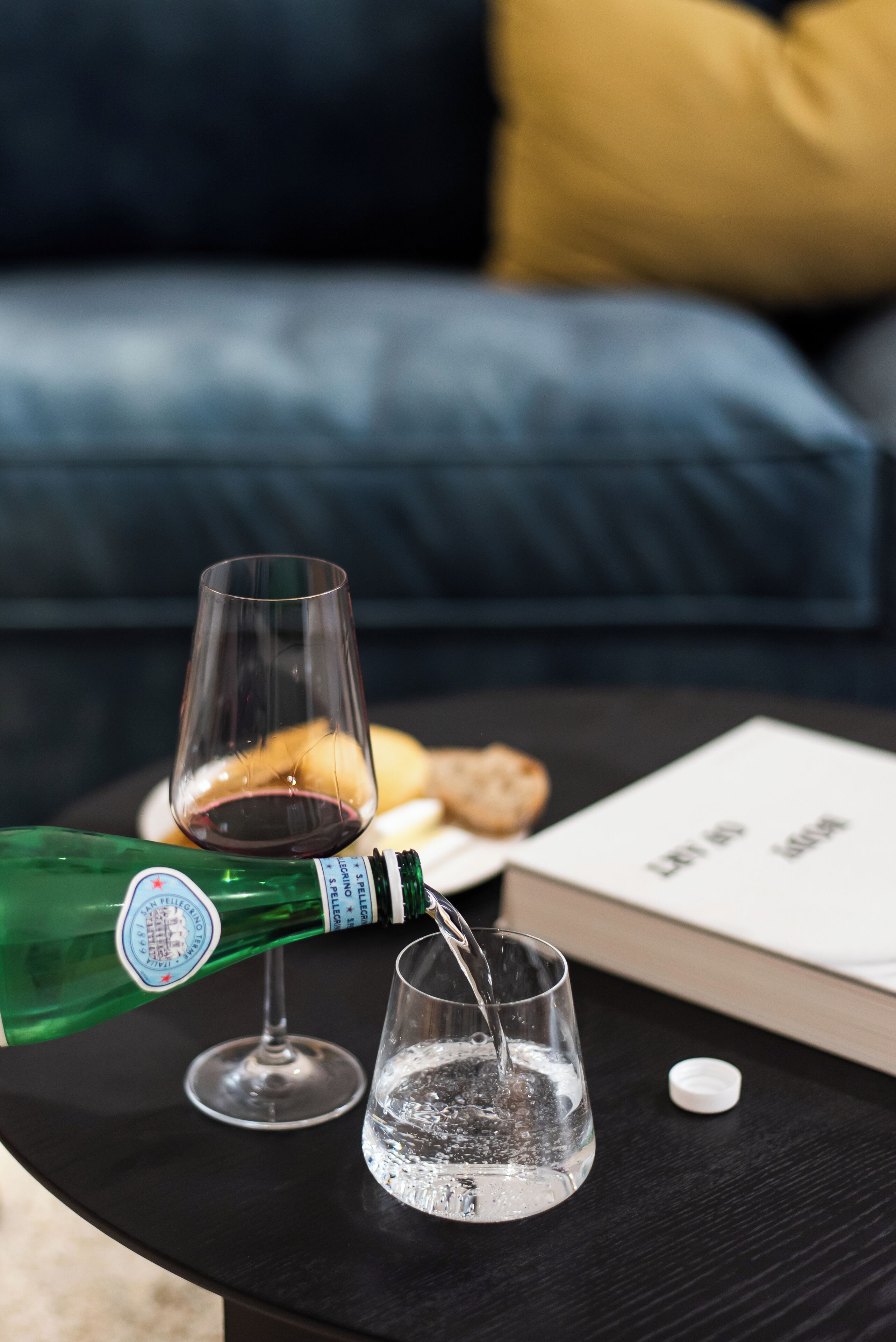 A bottle of champagne being poured into a glass on a black table, with a book and a corkscrew nearby.