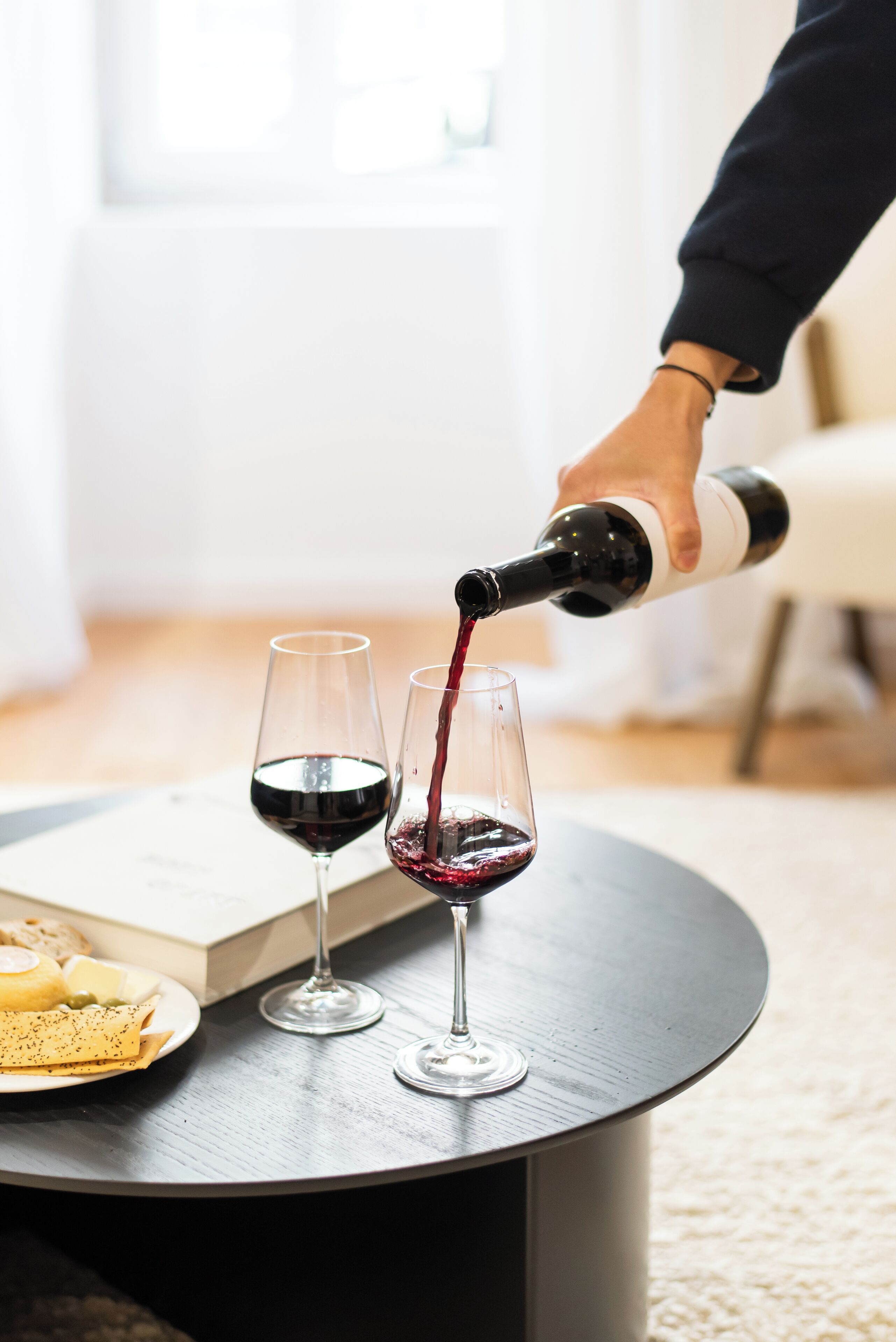 Eine Person gießt Wein in zwei Gläser auf einem runden Tisch, neben denen sich ein Buch und eine Käseplatte befinden.