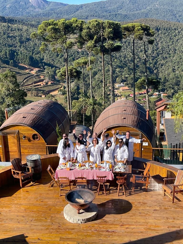Un grupo de personas con batas blancas reunidas alrededor de una mesa con mantel a cuadros en una terraza al aire libre con mobiliario de madera, rodeada por dos estructuras de forma abovedada con paredes de madera y un paisaje montañoso al fondo.