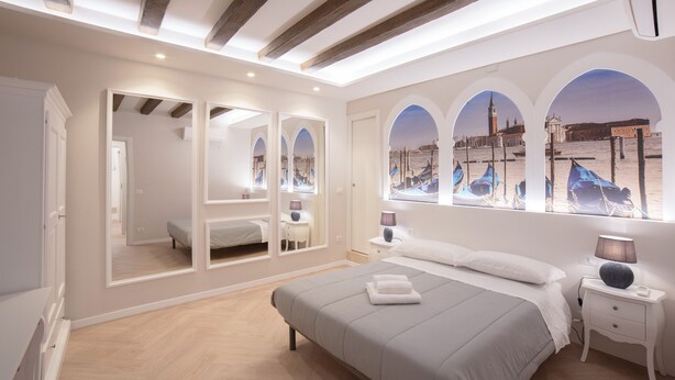 A modern bedroom with a large bed, a nightstand, a mirror, and a view of boats through arched windows. - Ca di Viola e Tobia (Venice)