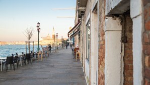 A waterfront promenade with tables and chairs, a brick building on the right, and a distant view of a tower. - Ca di Viola e Tobia (Venice)