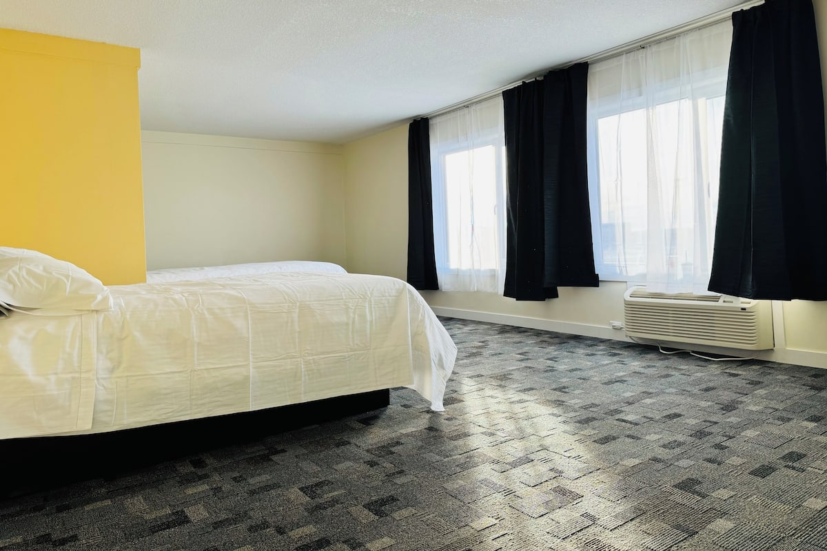 A hotel room with a bed, a window with black curtains, a ceiling light, and a wall heater.